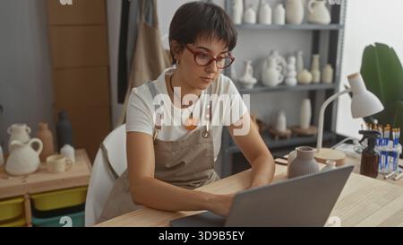 Young woman in apron and glasses types on laptop with both hands beside finished ceramic vases and tools on table in pottery studio; concentration. Stock Photo