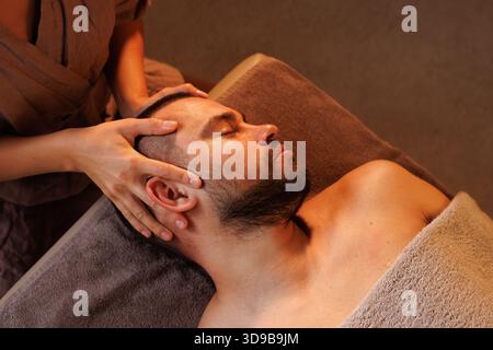 female massage therapist doing facial massage to man, close-up ear massage, in warm atmosphere of massage parlor, , massage concept Stock Photo