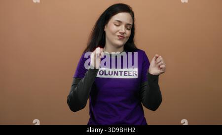 Woman dancing in purple volunteer shirt against orange background smiles joyfully, showcasing volunteerism, happiness, and positivity, with hands rais Stock Photo