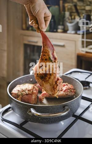 A person holds a marinated rabbit hind leg above a pot with other meat cooking inside. The kitchen has wooden cabinets and a warm atmosphere, indicati Stock Photo