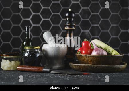 A variety of fresh vegetables such as tomatoes, onions, and greens sit on a dark kitchen counter alongside cooking tools Stock Photo
