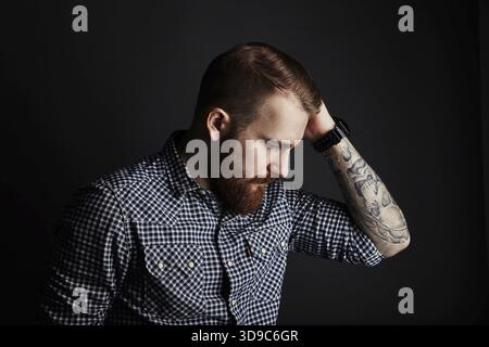 Thoughtful red bearded handsome stylish man with tattooes in blue checkered shirt, studio portrait on dark background Stock Photo