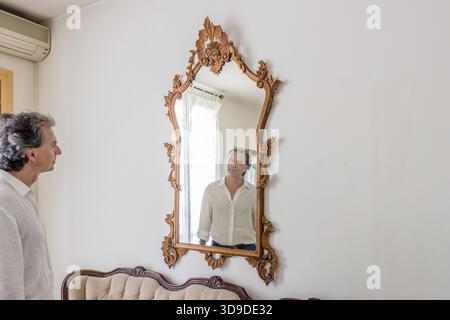 Sporty elegant middle-aged man with gray hair is reflected in a large mirror vintage in his living room Stock Photo