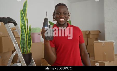 Man holding drill beside ladder and potted plant in building cluttered with moving boxes and stacked cartons, smiling broadly; home improvement joy. Stock Photo
