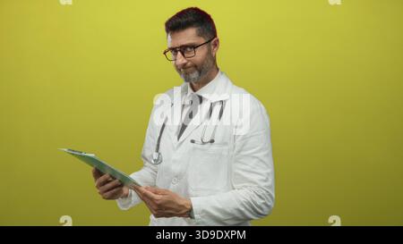 Man doctor wearing stethoscope reading clipboard and checking medical notes in studio with yellow backdrop; focused professionalism. Stock Photo