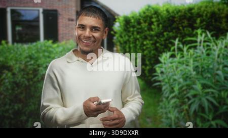 Man holding smartphone and smiling while looking down in front of a building near a house and hedges; quiet contentment. Stock Photo