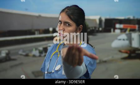 Young hispanic woman doctor beckons with hand and stethoscope at airport terminal beside airplane; authority. Stock Photo