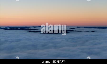 Aerial view of dark land formations emerging from a sea of white clouds under a gradient sky painted with soft hues of orange and pink., Miranda, Cali Stock Photo