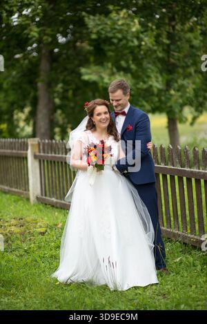 Young wedding couple embracing near a wooden fence in Latvia, sharing a warm and joyful wedding moment outdoors. Stock Photo