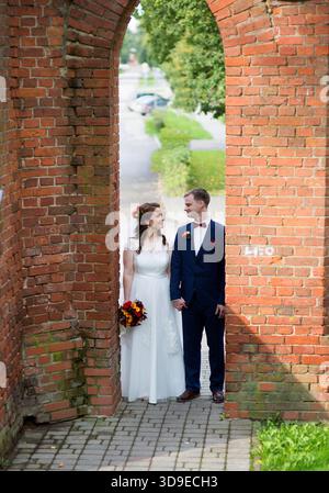 Young adult wedding couple holding hands between red brick walls in Latvia, smiling at each other with visible joy, formal attire and colorful bouquet Stock Photo