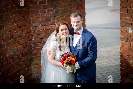 Young adult newlyweds embracing gently near a brick archway in Latvia, smiling with visible happiness, surrounded by soft daylight and festive wedding Stock Photo