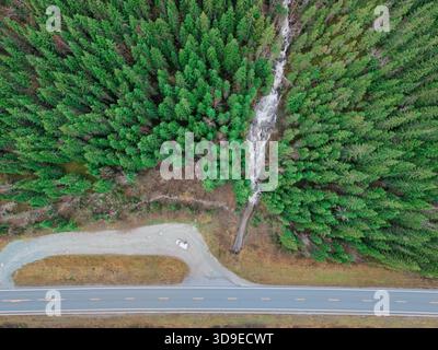 Aerial view of a road running parallel to a river cutting through a dense forest, painting a contrast between the grey asphalt and vibrant green canop Stock Photo