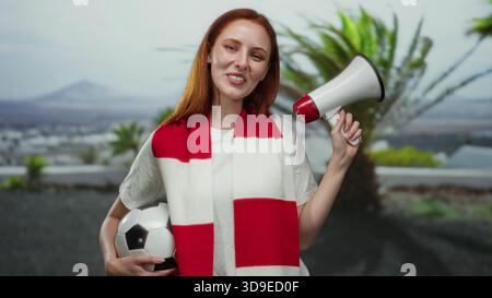 Woman smiling outdoors holding a megaphone and soccer ball, wearing a red and white scarf in a city street, embodying patriotic football enthusiasm. Stock Photo