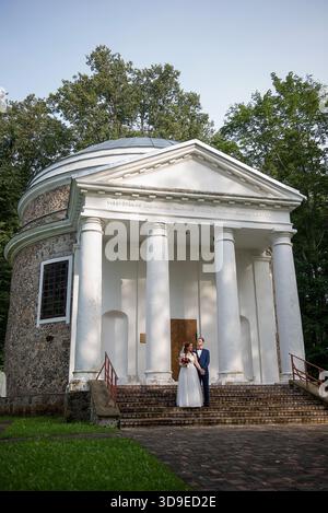 Middle aged bride and groom stand together on stone steps of an old chapel, holding hands calmly and posing in elegant wedding attire with bouquet and Stock Photo