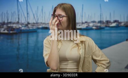 Blonde woman wearing glasses pinches nose beside boats on sunlit street pier over blue water; disgust. Stock Photo