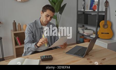Man writes notes on paper with a yellow pen at a wooden desk beside a laptop and calculator in a studio; focus productivity. Stock Photo