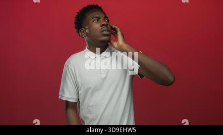 Man wearing white polo touches cheek then rests hand on chin and gazes upward in red studio; contemplation. Stock Photo