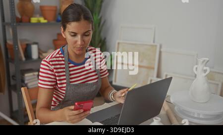 Woman holding red smartphone and writing with pen while using laptop in studio; creative concentration. Stock Photo