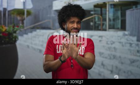 Lifeguard in red shirt makes rejecting hand gestures on city street lined with outdoor steps, expressing disapproval or surprise while smiling slightl Stock Photo