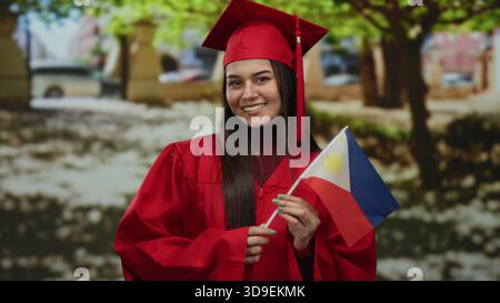 Woman in red graduation gown holding philippine flag in a park setting, symbolizing accomplishment, pride, and national identity. Stock Photo