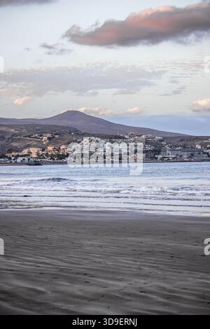 View of a serene beach with gentle waves kissing the shore, leading to a distant town nestled beneath a mountain under a pastel sky, Gran Canaria, Can Stock Photo