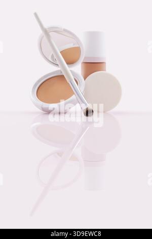 Opened compact powder, and a closed foundation in a pump bottle, soft brush and makeup puff. Cosmetics beauty products isolated on a glossy reflective Stock Photo