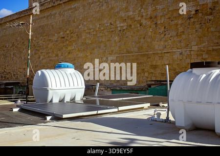 Modern white plastic water tanks and solar panels stand on a sunny rooftop against a fortress wall in Valletta, Malta. Stock Photo