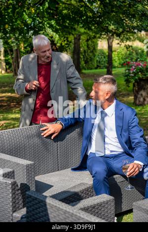 Candid scene of two adult men engaged in conversation during an outdoor formal event. One man is seated with a glass of wine, while the other stands n Stock Photo