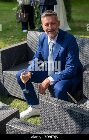 Portrait of a well-dressed mature man sitting on outdoor wicker furniture during a formal event. He is wearing a blue suit and holding a glass of whit Stock Photo