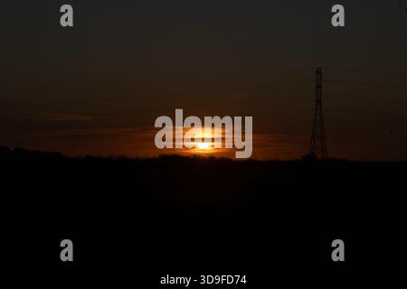 The lower half of the sun sets behind a dark line of trees, with a prominent electric pylon silhouette and power lines against an orange and brown sky Stock Photo