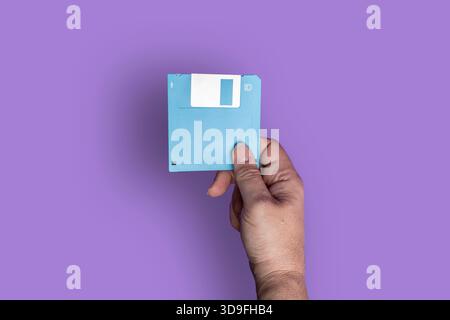 Close-up of a person’s hand holding a blue floppy disk against a purple background, symbolizing retro tech and data storage. Stock Photo