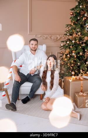 A couple shares a joyful moment in a beautifully decorated room for Christmas, posing beside a tall tree adorned with lights, ornaments, and surrounde Stock Photo