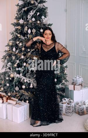 In a warm indoor setting, a woman in a black sparkling dress poses near a beautifully decorated Christmas tree. Stock Photo