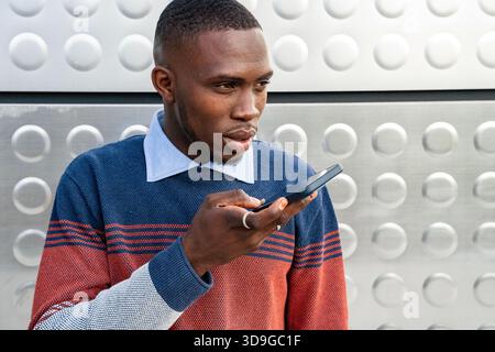 A man is engaged in a conversation using his smartphone in a modern urban setting. He looks focused, speaking clearly as he stands against a textured Stock Photo