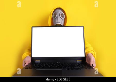 Individual in yellow hoodie and gas mask holding laptop with blank white screen on vibrant yellow background, copy space. Stock Photo