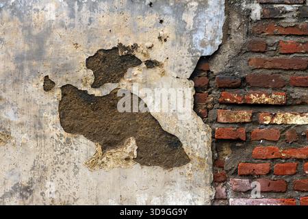 High-contrast texture split between severely damaged, peeling light grey plaster/stucco and a section of vibrant, exposed red brick. Large irregular h Stock Photo