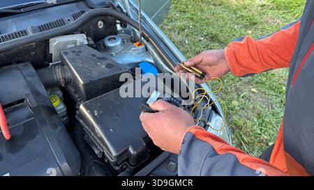 A Caucasian male mechanic in an orange and gray jacket adjusting a glow plug tester while holding glow plugs near the open engine bay of a silver car. Stock Photo