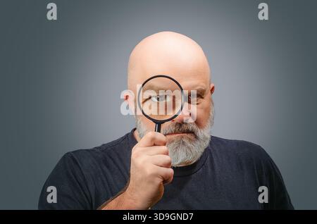 Bearded man looking through magnifying glass with big, enlarged, funny eye, searching, analyzing or investigating. Stock Photo