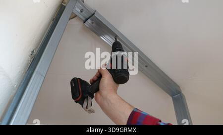 A hand holds a screwdriver and secures a metal structure to the ceiling, attaching a metal profile to a wall and ceiling surface Stock Photo