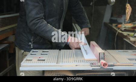 An impersonal carpenter works on a circular saw, cutting a narrow wooden plank lengthwise against the background of an equipped workshop Stock Photo