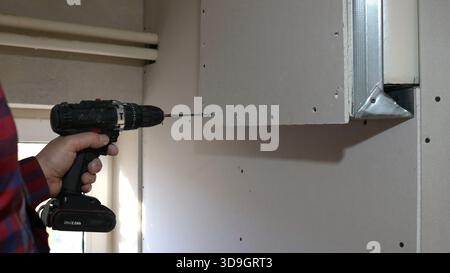 A hand with a screwdriver secures a panel to a profiled metal structure on a wall in a room against the backdrop of white water pipes and window Stock Photo