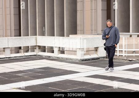 Middle-aged man standing on rooftop terrace holding smartphone and crossbody camera bag, copy space Stock Photo