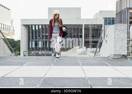 Female student walking down campus plaza carrying backpack wearing headphones with paper coffee cup Stock Photo