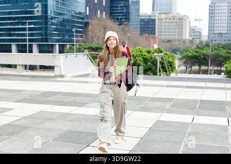 Female student walking across downtown plaza holding green and red notebooks with backpack Stock Photo