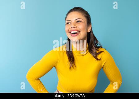Woman wearing yellow turtleneck sweater, smiling, posing hands on hips in studio with blue backdrop Stock Photo