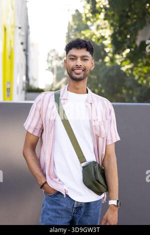 Indian man standing casually against low gray wall on sunny walkway with green crossbody bag Stock Photo