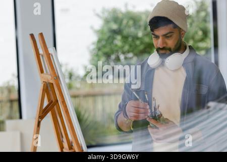 Indian man wearing headphones selecting paintbrushes at easel in art studio with canvas, copy space Stock Photo