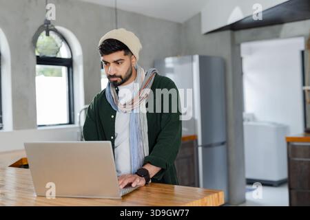 Indian man typing on silver laptop at kitchen island wearing beanie and scarf beside refrigerator Stock Photo