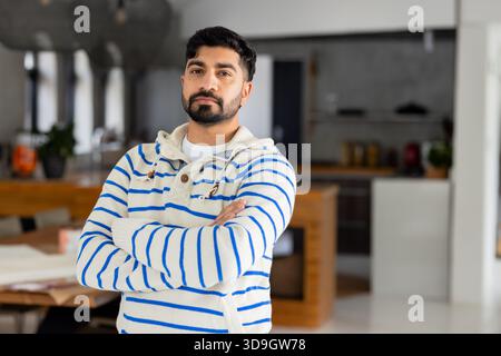Indian man standing with arms crossed in modern kitchen beside table with papers, mug and plant Stock Photo