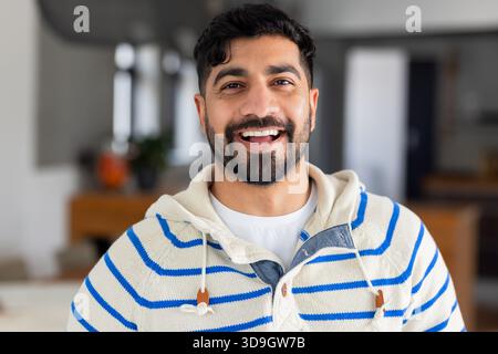 Indian man smiling and leaning on wood countertop in kitchen wearing striped hoodie Stock Photo